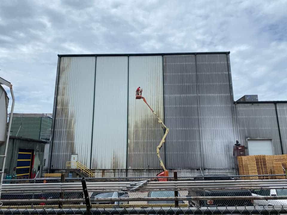 A person operates a cherry picker to clean the exterior wall of a large industrial building under a cloudy sky.